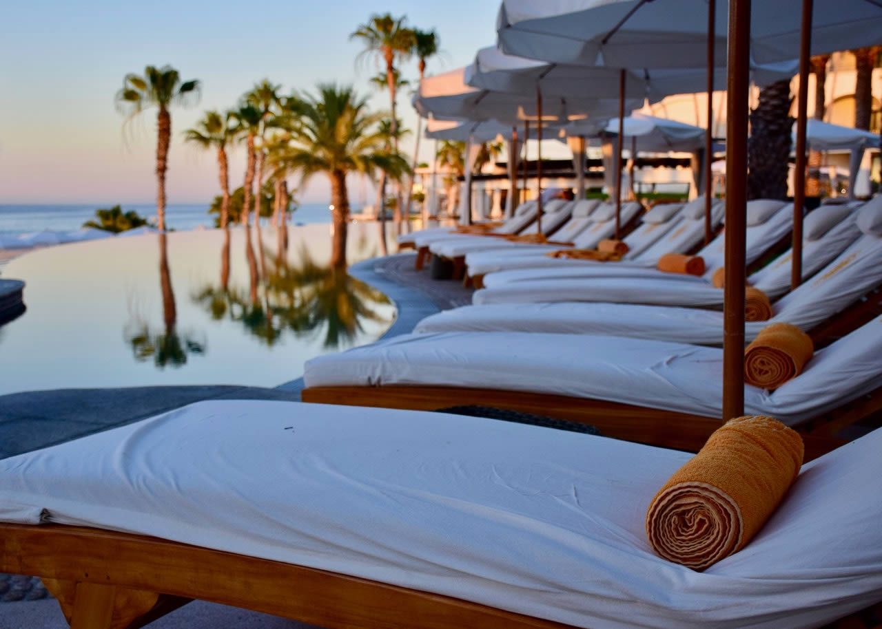 Sun loungers with white cushions and rolled towels beside a resort swimming pool at sunset, with palm trees and sea in the background