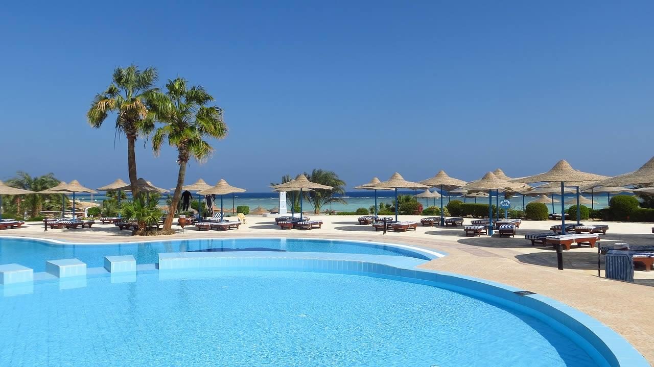 Outdoor resort swimming pool with sun loungers and straw parasols, palm trees and sea view under a clear blue sky