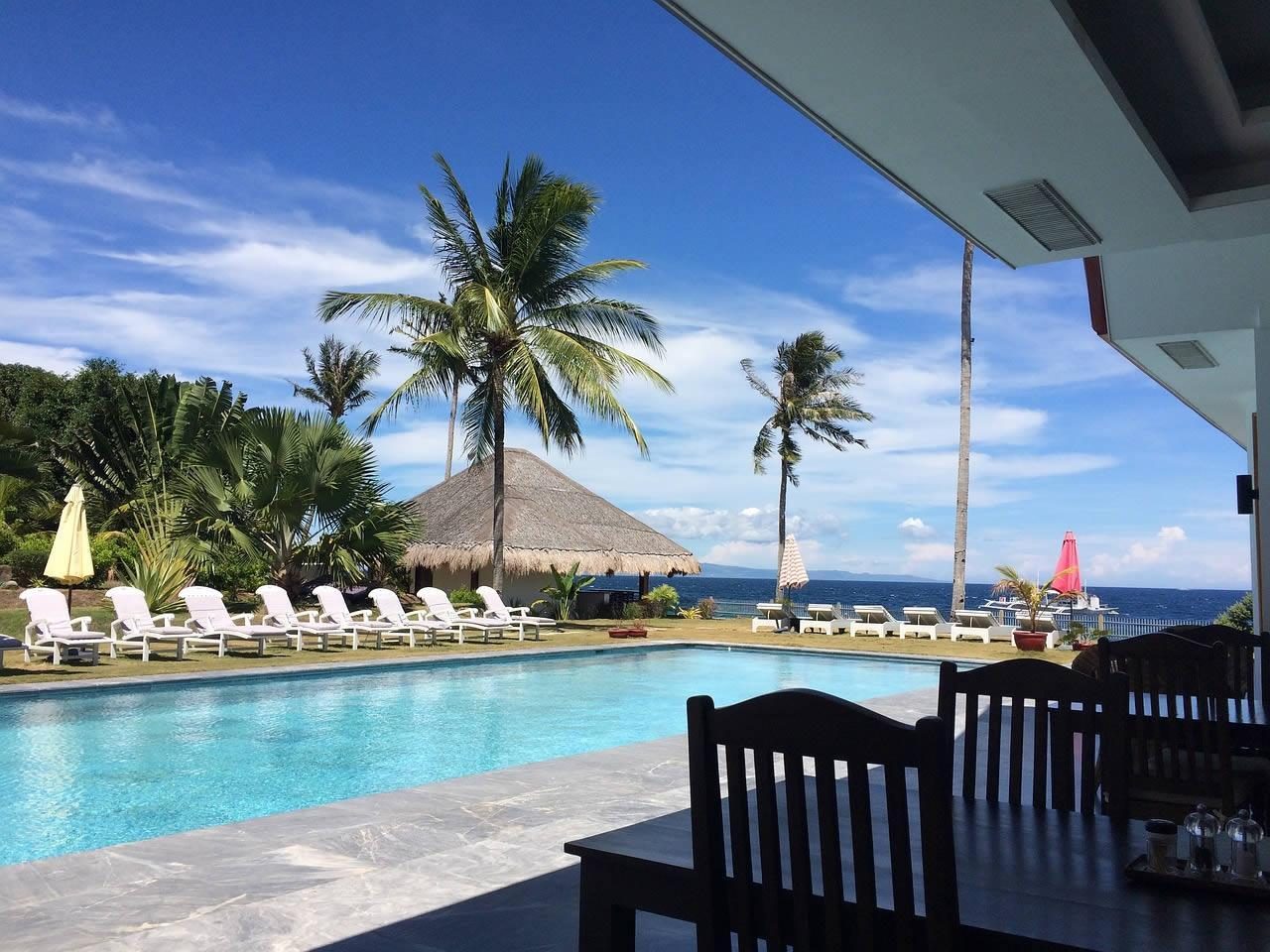 Resort swimming pool with sun loungers, palm trees and a thatched hut on a terrace overlooking the sea under a blue sky