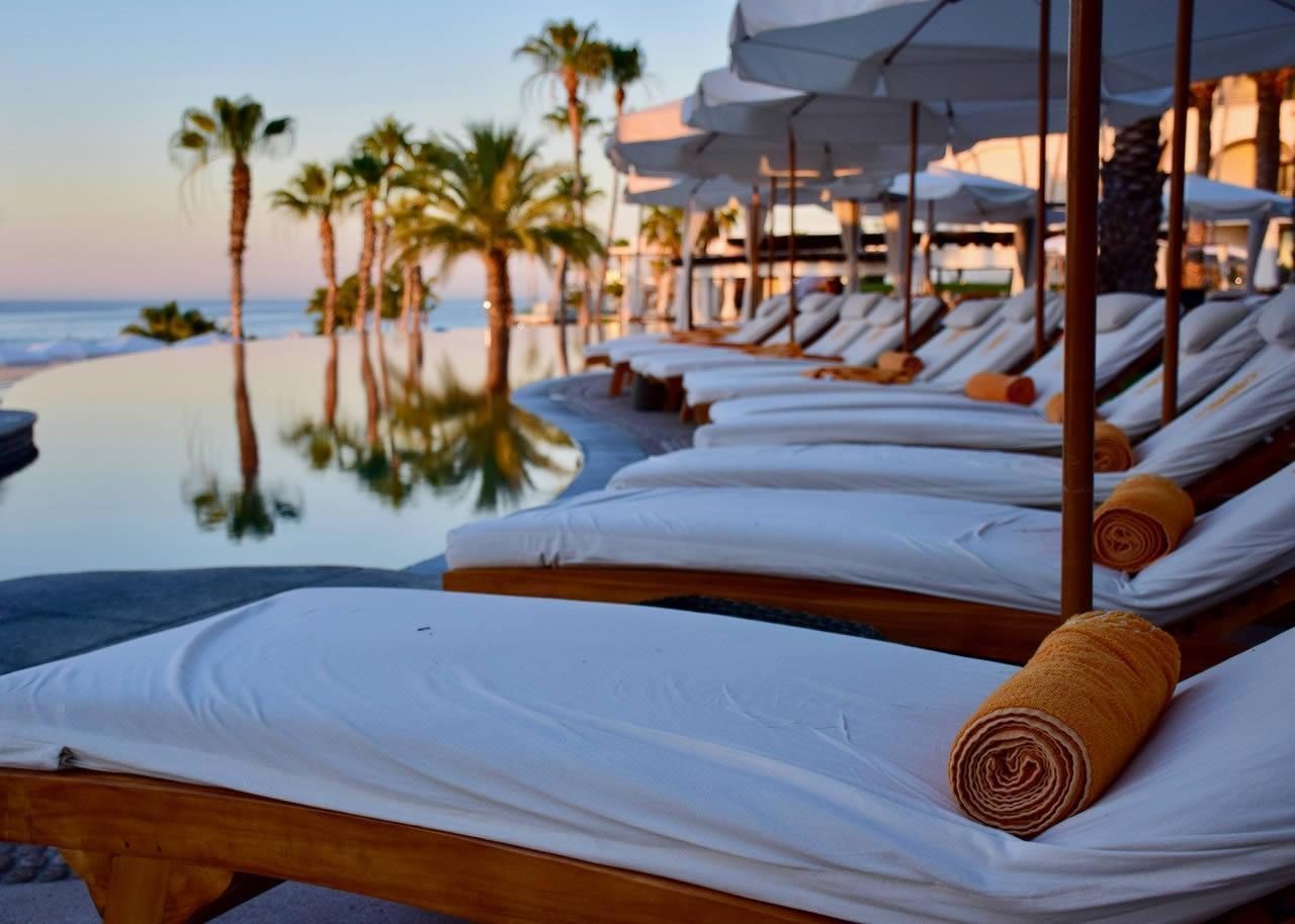 Sun loungers with rolled towels under white parasols beside a palm-lined resort pool overlooking the sea at sunset