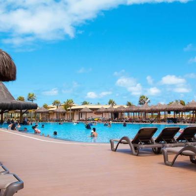 Sun loungers around a large resort swimming pool with thatched parasols and people swimming under a bright blue sky