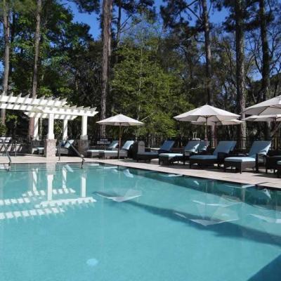 Outdoor resort swimming pool with sun loungers and parasols beside a white pergola, surrounded by tall trees