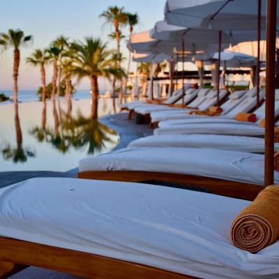 Sun loungers with rolled towels under white parasols beside a palm-lined resort pool overlooking the sea at sunset
