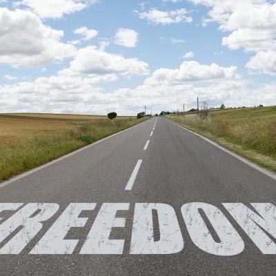 Open road through countryside with the word “FREEDOM” painted in large white letters on the tarmac under a cloudy sky